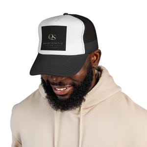 Op de afbeelding: Zwarte en witte truckerpet met een vierkant logo met de tekst "QUIETSTATIC CO." in witte letters op een zwarte achtergrond. De pet heeft een zwarte klep en mesh achterkant, en een wit voorpaneel.