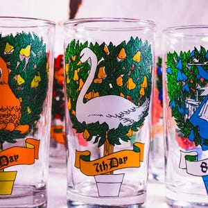 May include: Three clear glass tumblers with colorful illustrations of the 6th, 7th, and 8th days of Christmas. Each tumbler features a different scene with a goose, a swan, and a woman in a blue dress.