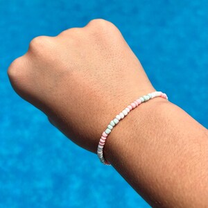 May include: A pastel beaded bracelet featuring a pattern of white, pink, and light green beads. The bracelet is worn on a person's arm, with a blue background.