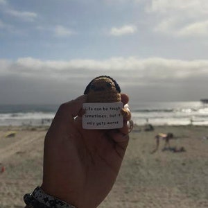 Op de afbeelding: Een klein gehaakt speelgoedje dat een bordje vasthoudt met de tekst "Het leven kan soms zwaar zijn, maar het wordt alleen maar erger". Het speelgoedje wordt vastgehouden door een hand met een armband om de pols. De achtergrond is een strand met de zee in de verte.