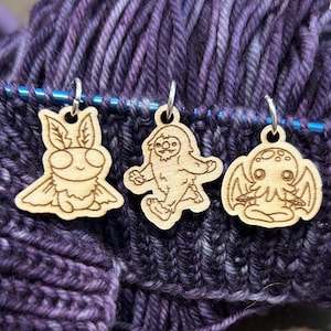 May include: Three wooden stitch markers with silver hoops. The markers feature a moth, a Bigfoot, and a Cthulhu design. The markers are on knitting needles with purple and blue yarn in the background.