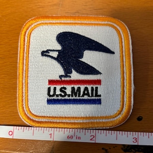 May include: A white embroidered patch with a yellow border. The patch features a blue eagle with red, white, and blue stripes below it and the text "U.S. MAIL".