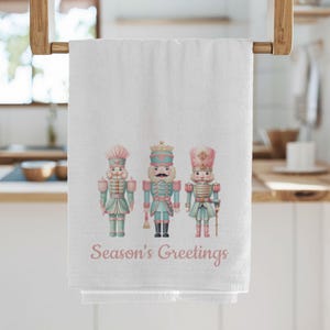 Op de afbeelding: Witte keukendoek met drie pastelkleurige notenkrakersoldaten. De tekst "Season's Greetings" staat in roze letters. De handdoek hangt aan een houten handdoekenrek.