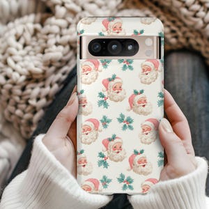 May include: A phone case featuring a repeating pattern of Santa Claus faces with pink hats and white beards, interspersed with sprigs of holly and berries. The case has a cream-colored background and is held in two hands.