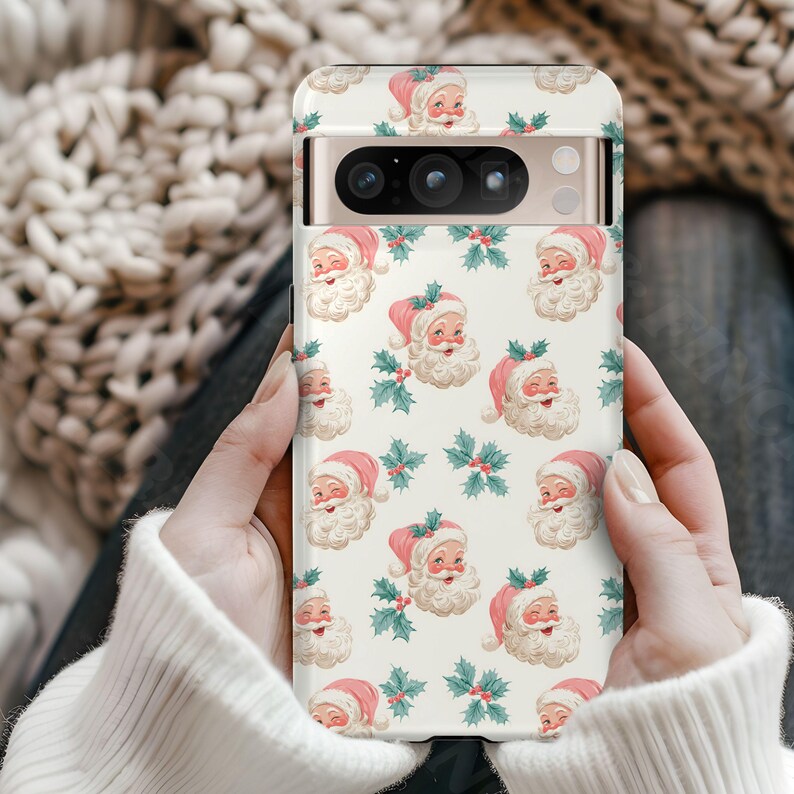 May include: A phone case featuring a repeating pattern of Santa Claus faces with pink hats and white beards, interspersed with sprigs of holly and berries. The case has a cream-colored background and is held in two hands.