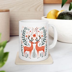 May include: White ceramic mug with a colorful design of two deer with antlers facing each other in a floral pattern. The deer are orange and the flowers are red, pink, and green.