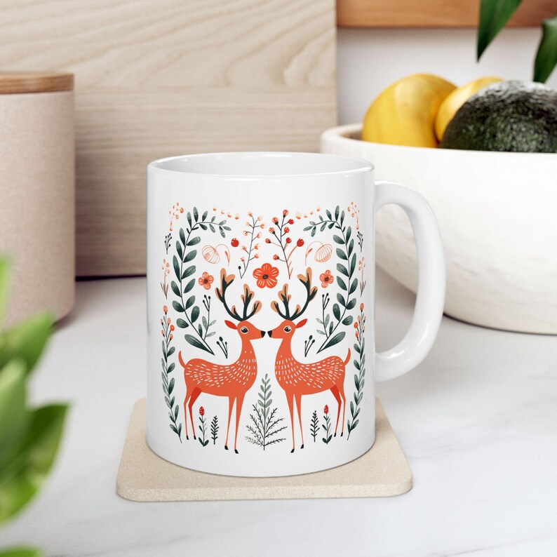 May include: White ceramic mug with a colorful design of two deer with antlers facing each other in a floral pattern. The deer are orange and the flowers are red, pink, and green.