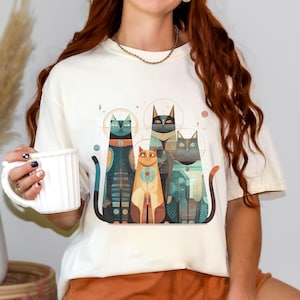 May include: Cream-colored t-shirt featuring a stylized illustration of five cats. The cats are depicted in various colors, including teal, orange, and gray, with geometric patterns and celestial accents. The design has a retro, artistic style.