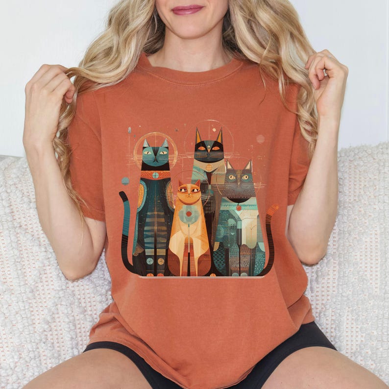 May include: Burnt orange t-shirt featuring a graphic of five stylized cats with geometric patterns and halos. The cats are in shades of blue, orange, and gray. The shirt is a crew neck and has short sleeves.