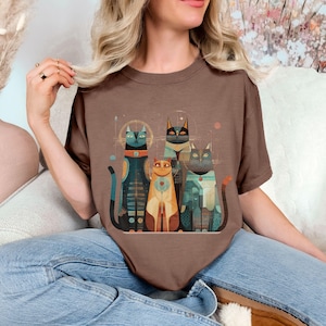 May include: A brown t-shirt featuring a stylized illustration of four cats. The cats are depicted in various colors, including teal, orange, and black, with geometric patterns and details. The design has a retro, artistic style.