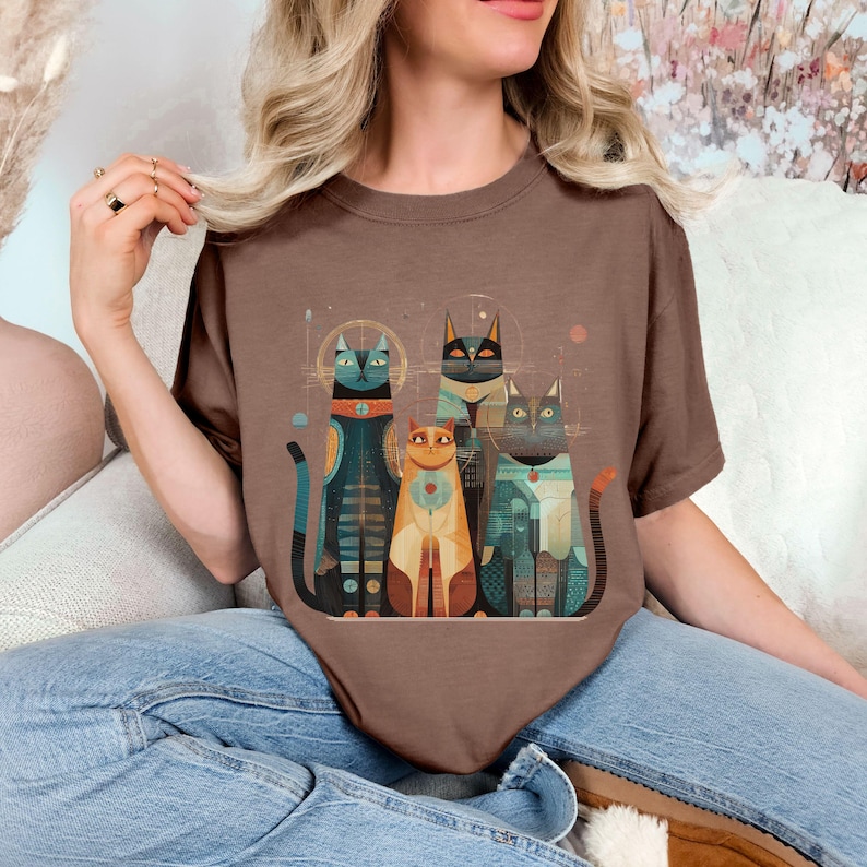 May include: A brown t-shirt featuring a stylized illustration of four cats. The cats are depicted in various colors, including teal, orange, and black, with geometric patterns and details. The design has a retro, artistic style.