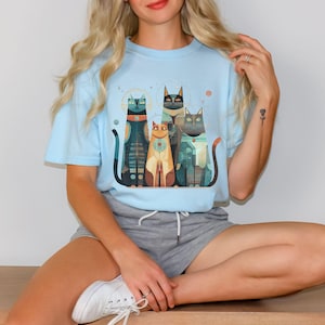 May include: Light blue t-shirt featuring a colorful graphic of four stylized cats. The cats have unique patterns and colors, including black, orange, and teal. The design has a geometric and artistic style, with a celestial theme.