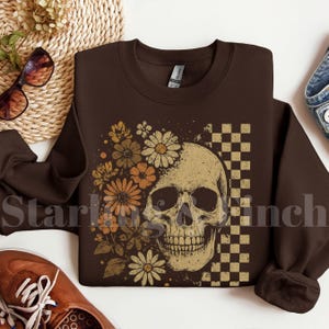 Puede incluir: Sudadera marrón oscuro con un diseño de calavera, flores y un patrón de cuadros. La calavera es beige, las flores son naranjas y beige, y el patrón de cuadros es beige y marrón. Prenda de vestir informal.
