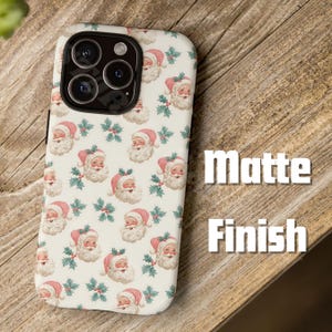May include: A phone case with a matte finish, featuring a repeating pattern of Santa Claus faces and holly sprigs on a cream background. The case is designed to fit a smartphone, with cutouts for the camera and other features.