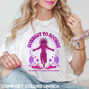 May include: White t-shirt with a pink and purple graphic design featuring a figure with an eye in the center of its head. The graphic design includes the text "Connect to Source" and "The Gateway to Higher Consciousness".