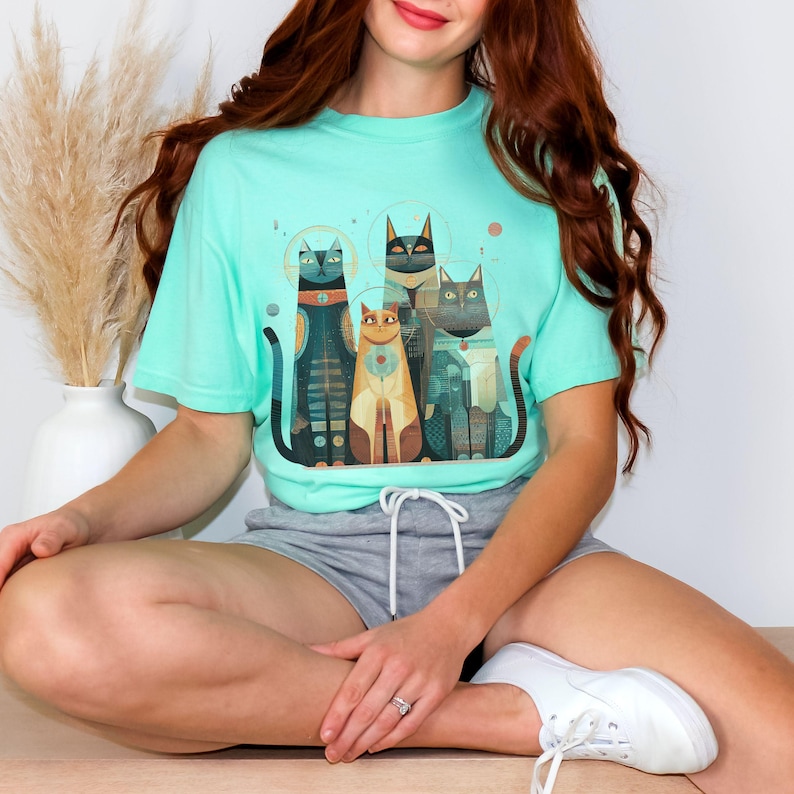 May include: A light teal t-shirt featuring a graphic of five stylized cats in various colors and patterns. The cats have geometric designs and are arranged in a row. The shirt is worn by a person.