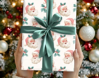 May include: A wrapped gift with a green satin ribbon and bow. The wrapping paper features a repeating pattern of Santa Claus faces with pink hats and holly sprigs on a cream background. The gift is held in front of a Christmas tree.