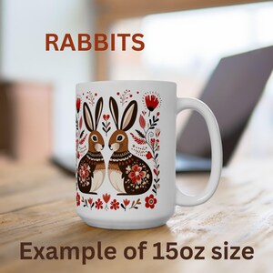 May include: White ceramic mug with a floral design featuring two brown rabbits and the word "RABBITS" printed in brown at the top. The mug is 15 ounces.