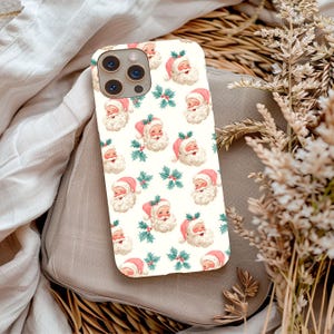 May include: A phone case with a repeating pattern of Santa Claus faces and holly sprigs on a cream background. Santa wears a pink hat and has a white beard. The case has a camera cutout and is a holiday-themed accessory.