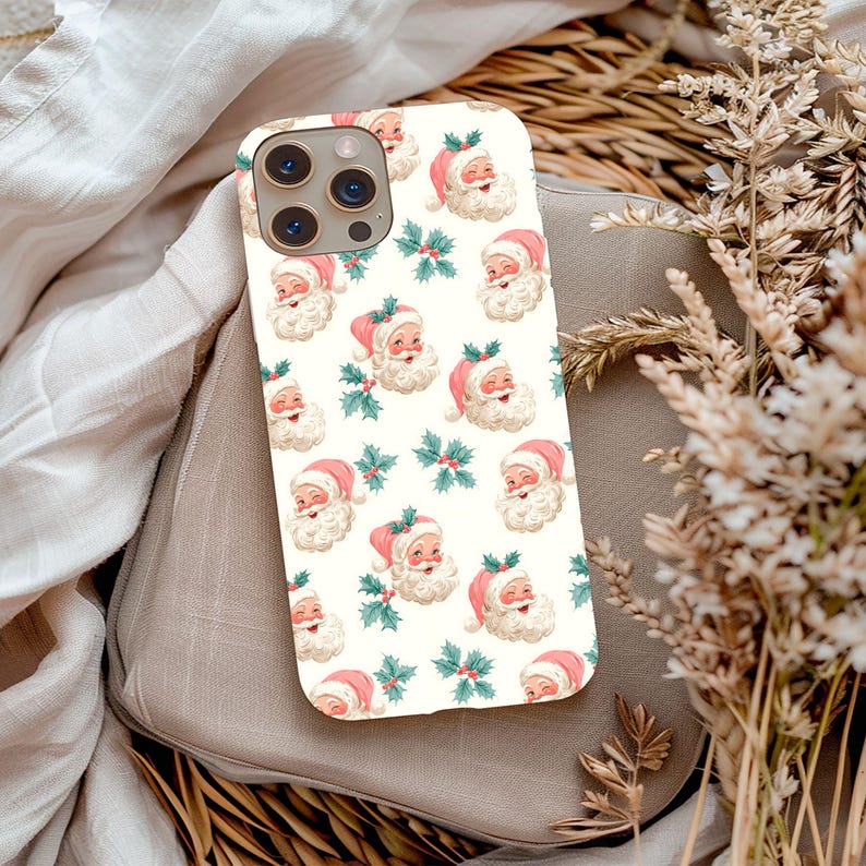 May include: A phone case with a repeating pattern of Santa Claus faces and holly sprigs on a cream background. Santa wears a pink hat and has a white beard. The case has a camera cutout and is a holiday-themed accessory.