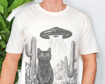 May include: Cream-colored t-shirt featuring a black and white graphic of a cat being abducted by a UFO above a desert landscape with cacti. The cat has wide eyes and an open mouth. The UFO is emitting a beam of light.