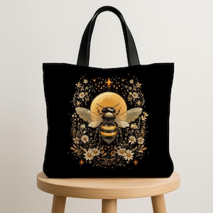 Pode incluir: Bolsa tote preta com uma ilustração de abelha, uma lua amarela e detalhes florais. A abelha tem listras pretas e amarelas e asas. O design é emoldurado por flores brancas e douradas. A bolsa tem alças pretas.