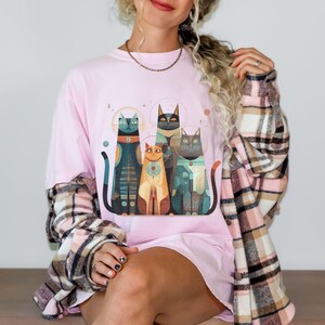 May include: A light pink t-shirt featuring a graphic of four stylized cats in teal, orange, and black. The cats have geometric patterns and halos. The shirt is worn with a plaid flannel shirt.