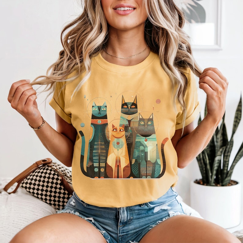 May include: Mustard yellow t-shirt featuring a graphic of four stylized cats in space suits. The cats are in shades of teal, orange, and black. The t-shirt has short sleeves and a crew neck.
