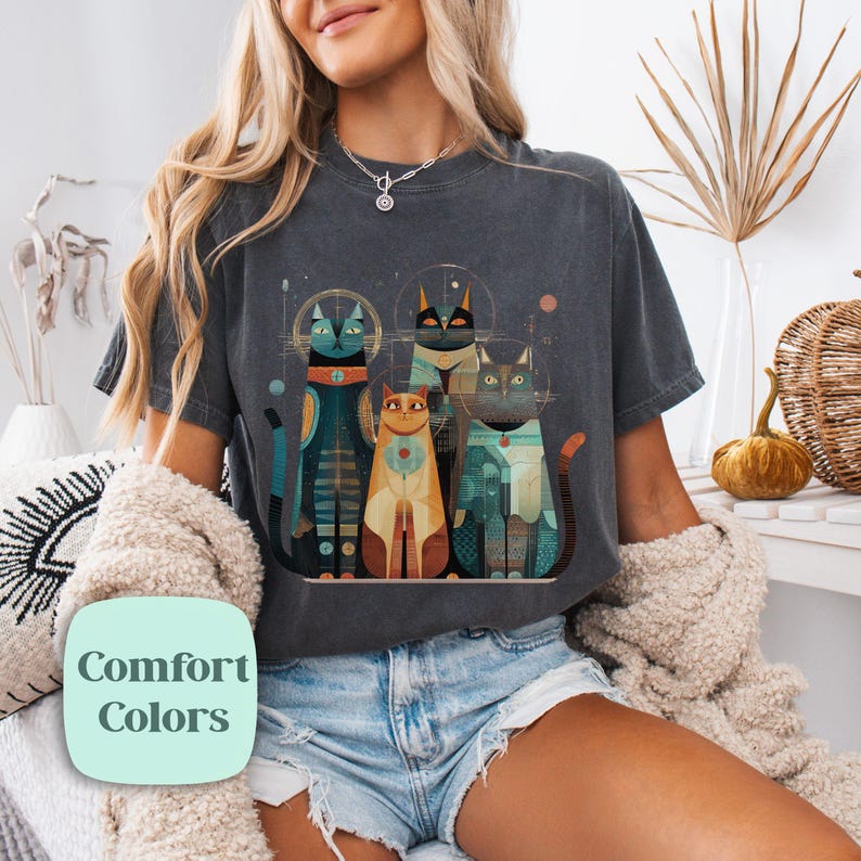 May include: Dark gray t-shirt featuring a stylized illustration of four cats with geometric designs and halos. The cats are in shades of blue, orange, and teal. The text "Comfort Colors" is in a light green box.