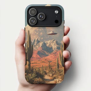 May include: A phone case featuring a desert landscape with cacti, mountains, and a UFO in the sky. The case has a blue and black border and a vintage, artistic style. The design evokes a sense of mystery and adventure.