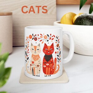 May include: White ceramic mug with a colorful illustration of two cats with floral accents. The text "CATS" is printed in bold letters above the image.
