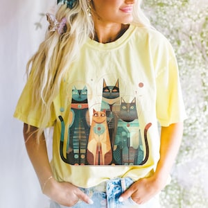 May include: Yellow t-shirt featuring a stylized illustration of four cats with geometric patterns in shades of teal, orange, and black. The cats have halos and are arranged in a row. The shirt is a light yellow color.