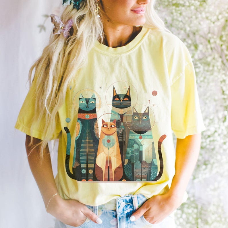 May include: Yellow t-shirt featuring a stylized illustration of four cats with geometric patterns in shades of teal, orange, and black. The cats have halos and are arranged in a row. The shirt is a light yellow color.