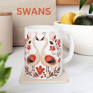 May include: White ceramic mug with a design of two swans facing each other in a heart shape. The swans are surrounded by red and brown floral designs. The text "SWANS" is printed in orange at the top of the mug.