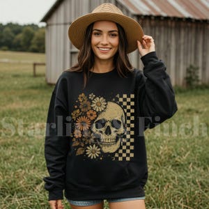 Puede incluir: Sudadera negra con un diseño de calavera, elementos florales y a cuadros. La calavera es beige con cuencas de ojos y nariz negras. Las flores son en tonos marrones y amarillos. El patrón de cuadros es negro y beige.