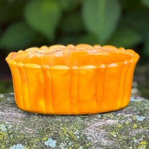 May include: An orange glass bowl with a scalloped rim sits on a mossy rock. The bowl has vertical ridges and a translucent quality, catching the light. Green foliage and pink flowers are blurred in the background, creating a natural setting.