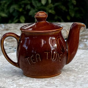 May include: A brown ceramic teapot with a lid and a handle. The teapot has the words "Tea Time" written on the side.