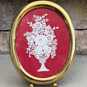 May include: Oval gold-toned frame with a red velvet background. A white lace floral design is centered within the frame. The frame has small ball feet for display.