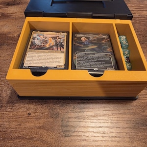 May include: A black and yellow plastic card storage box with a hinged lid and handle. The interior features three compartments containing two trading cards, Power Pack and Invisible Woman, and a small stack of translucent green dice.