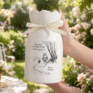 May include: A white ceramic vase with a scalloped top and a cream-colored ribbon tied around the neck. The vase features a black and white illustration of Winnie the Pooh and Piglet, with text that reads "Never forget the difference you make Mrs. Sophia" and "Thank you Jessica."