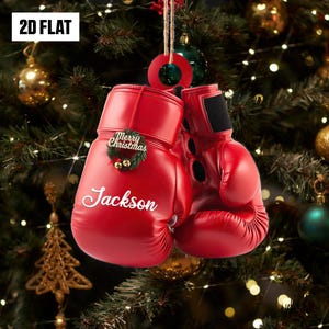 May include: A pair of red boxing glove Christmas ornaments. One glove has the name "Jackson" in white script and a "Merry Christmas" wreath. The ornaments hang from a string against a Christmas tree background.
