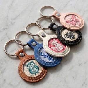 May include: Five circular keychains with metal rings, each featuring a leather-like border in tan, blue, white, black, or light pink. Each keychain contains a central fabric inlay embroidered with a different animal or insect: an owl, deer, fox, bee, or butterfly.