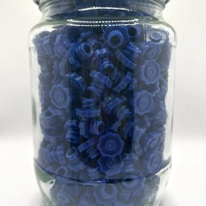 May include: A clear glass jar filled with numerous blue bottle stoppers. The stoppers have a flower-like design and are tightly packed inside the jar. The jar has a white lid.