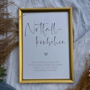 May include: A gold-framed sign with the text "Notfall-Körbchen" and a heart symbol. The sign also includes the text "FÜR NOTFÄLLE ALLER ART STEHT DIESES KORBCHEN PARAT. GREIFT ZU UND FEIERT ENTSPANNT WEITER!"
