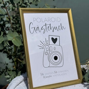Könnte beinhalten: Ein goldfarbener Rahmen mit dem Text "POLAROID Gästebuch" in schwarzer Kursivschrift. Unter dem Text befindet sich eine Schwarzweißillustration einer Polaroidkamera mit einem Herzen im Foto. Unter der Illustration steht der Text "Foto knipsen, Foto einkleben, Wünsche hinterlassen".