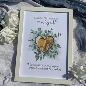Puede incluir: Un marco con texto en alemán que dice "Herzlichen Glückwunsch zur Hochzeit" y "Die schönsten Erinnerungen sammelt man immer zu zweit." El diseño en forma de corazón presenta un billete de euro rodeado de hojas verdes. El marco es de color beige claro.