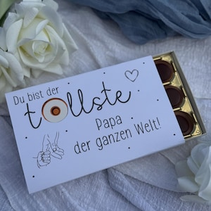 May include: A white box of chocolates with German text, including "Du bist der tollste Papa der ganzen Welt!" The box is open, showing several round chocolates. The image also features white roses and blue fabric.
