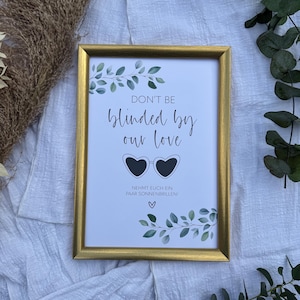 Könnte beinhalten: Ein goldfarbener Rahmen mit einem weißen Hintergrund und grünem Laub. Das Schild lautet "Don't be blinded by our love" mit einer Herzform-Sonnenbrille und dem Text "Nehmt euch ein paar Sonnenbrillen!"
