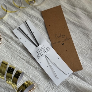 Könnte beinhalten: Eine weiße Karte mit dem Text "HAPPY NEW YEAR" und schwarzen Punkten, die zwei silberne Wunderkerzen hält. Daneben eine braune Karte mit "Frohes neues Jahr". Gold- und weiße Bänder und Perlen sind verstreut.