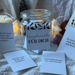 Può includere: Un barattolo di vetro trasparente con coperchio, pieno di piccoli biglietti bianchi. Il barattolo ha un'etichetta con la scritta "Komplimente FÜR DICH". Diverse carte con testo in tedesco sono sparse.